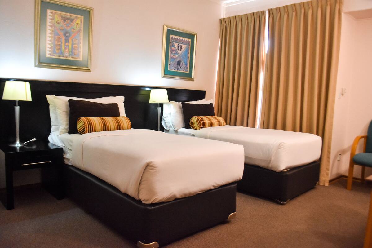 A hotel room with two beds, each with a black headboard, white bedding, and a lamp on a bedside table. There are two framed pictures on the wall and a window with curtains.