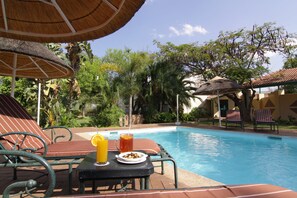 Un área junto a la piscina con sillones de descanso, una mesa con una bebida y un plato de comida, y una sombrilla de paja.