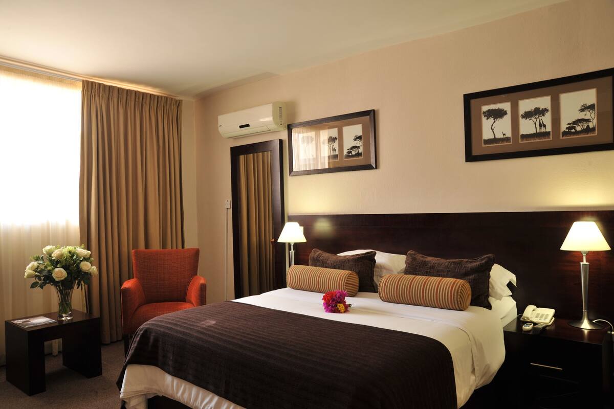 A hotel room with a large bed, two bedside lamps, a nightstand with a telephone, a chair, a vase with flowers, and a framed picture on the wall.