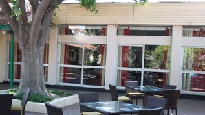 Un espace extérieur aménagé avec des chaises et des tables en osier, un grand arbre et un bâtiment doté de grandes fenêtres et de rideaux rouges.