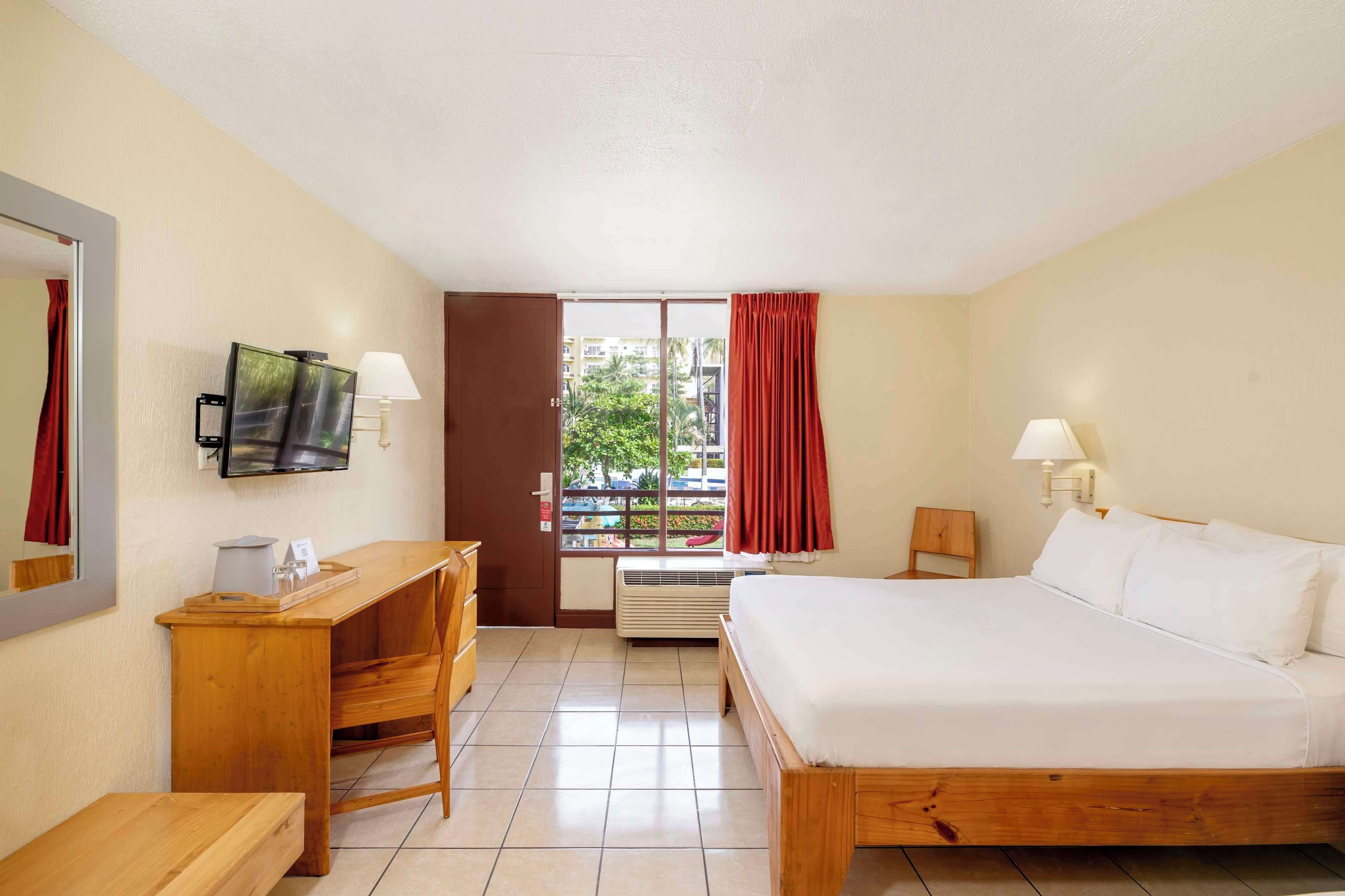 Una habitación de hotel con cama, escritorio, silla, televisor, ventana con cortinas y aire acondicionado.