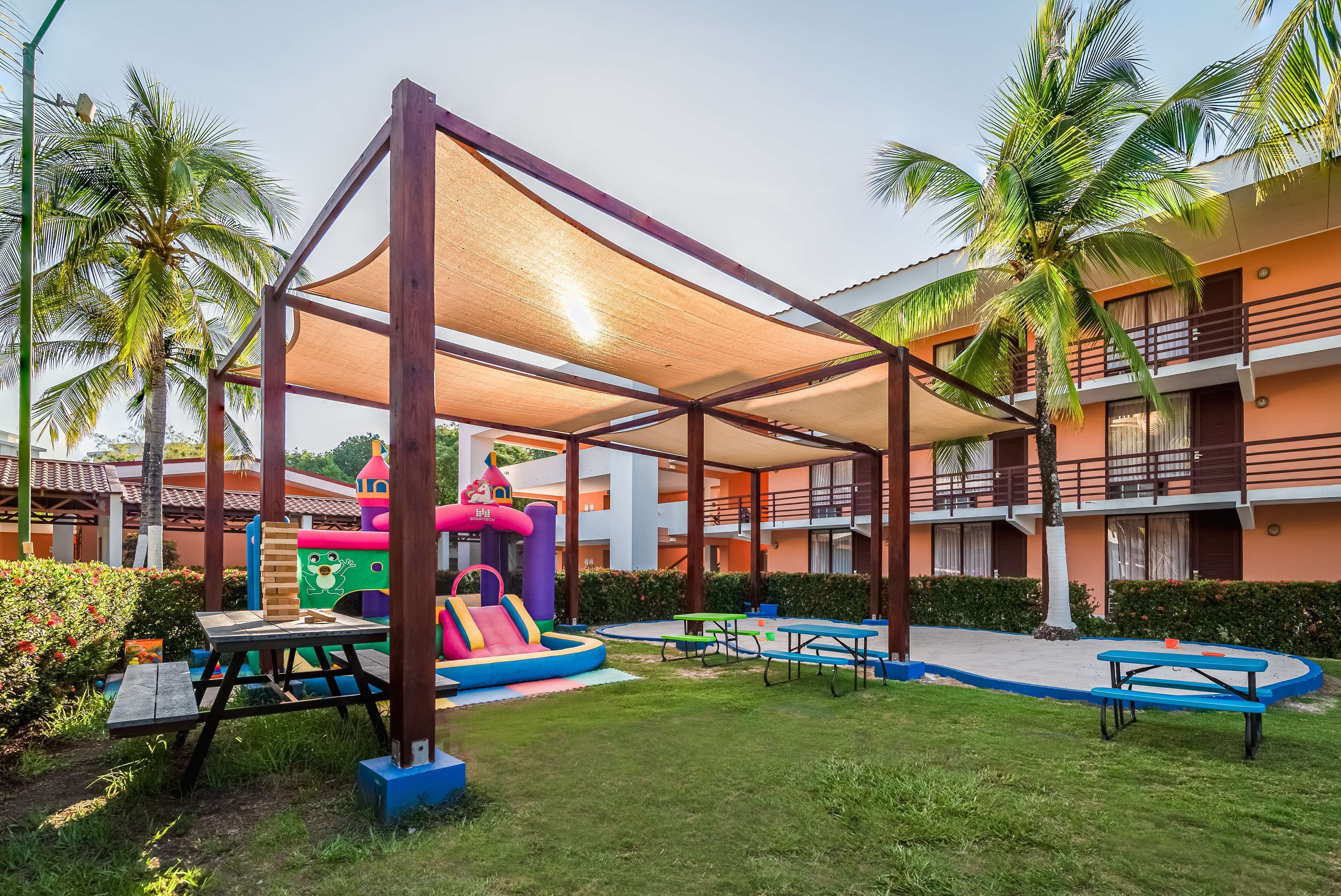 A children's play area with a slide and inflatable structures, surrounded by palm trees and a covered seating area.