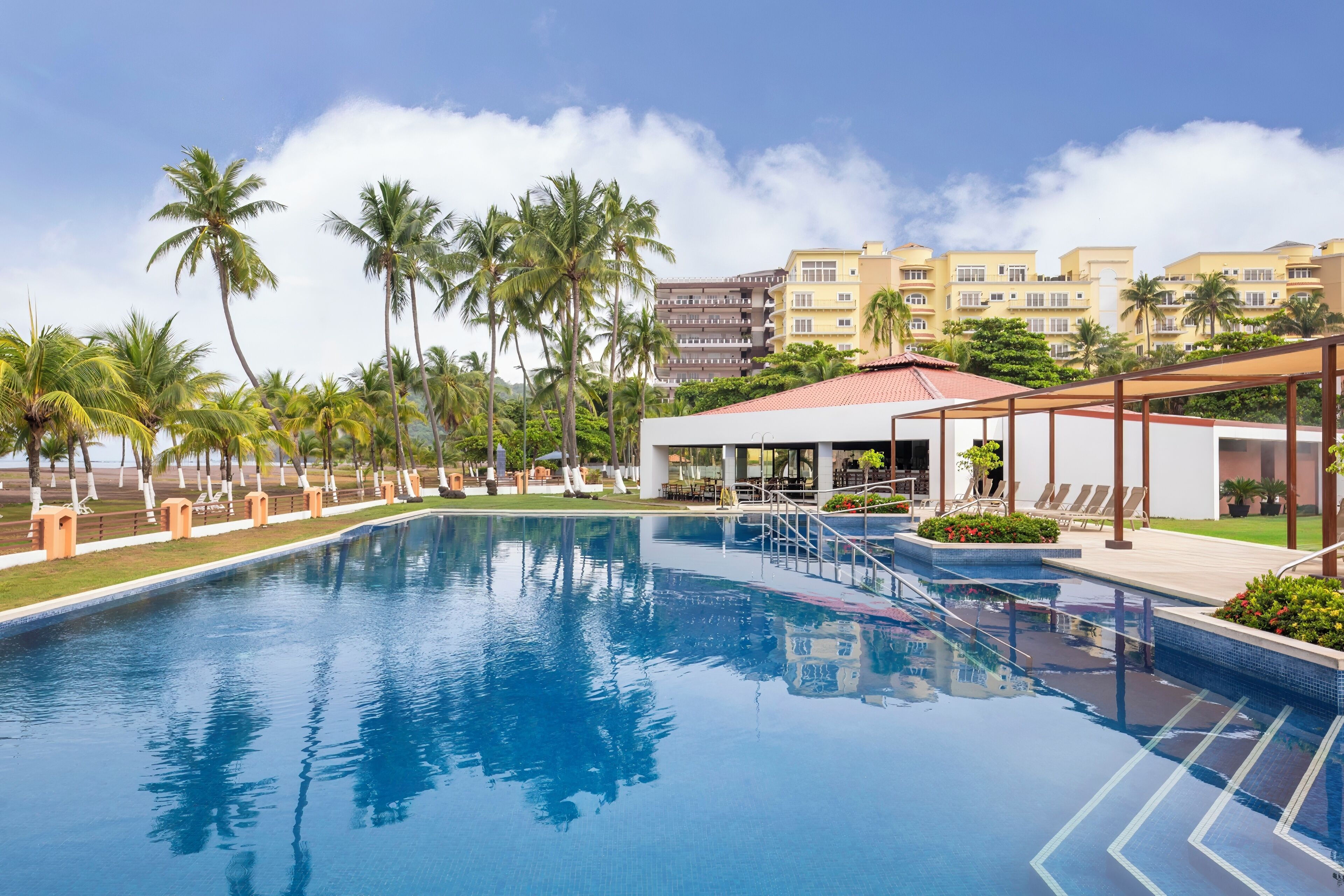 A swimming pool with lounge chairs and palm trees, surrounded by hotel buildings.
