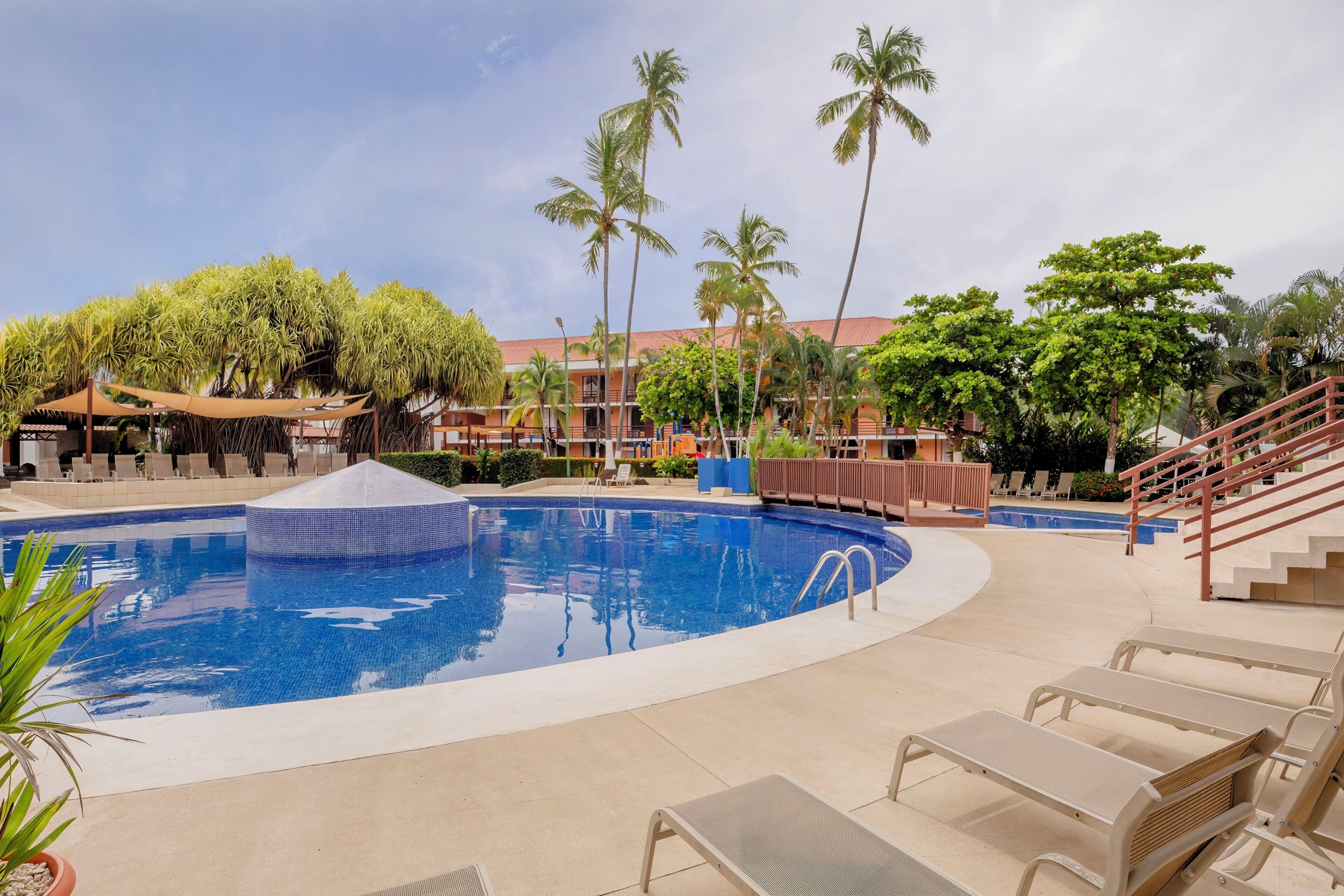 A pool area with lounge chairs, palm trees, and a building with a red roof.