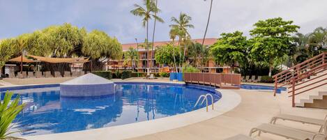 A pool area with lounge chairs, palm trees, and a building with a red roof.
