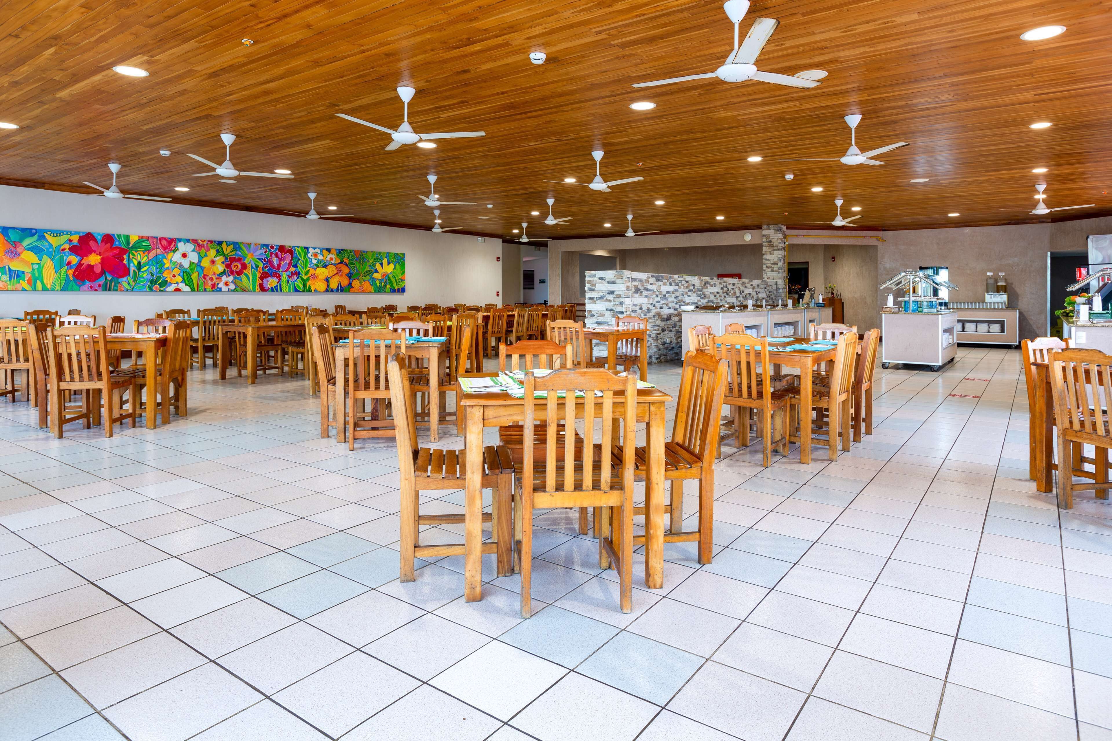 A spacious dining area with wooden tables and chairs, ceiling fans, and a colorful mural on the wall.