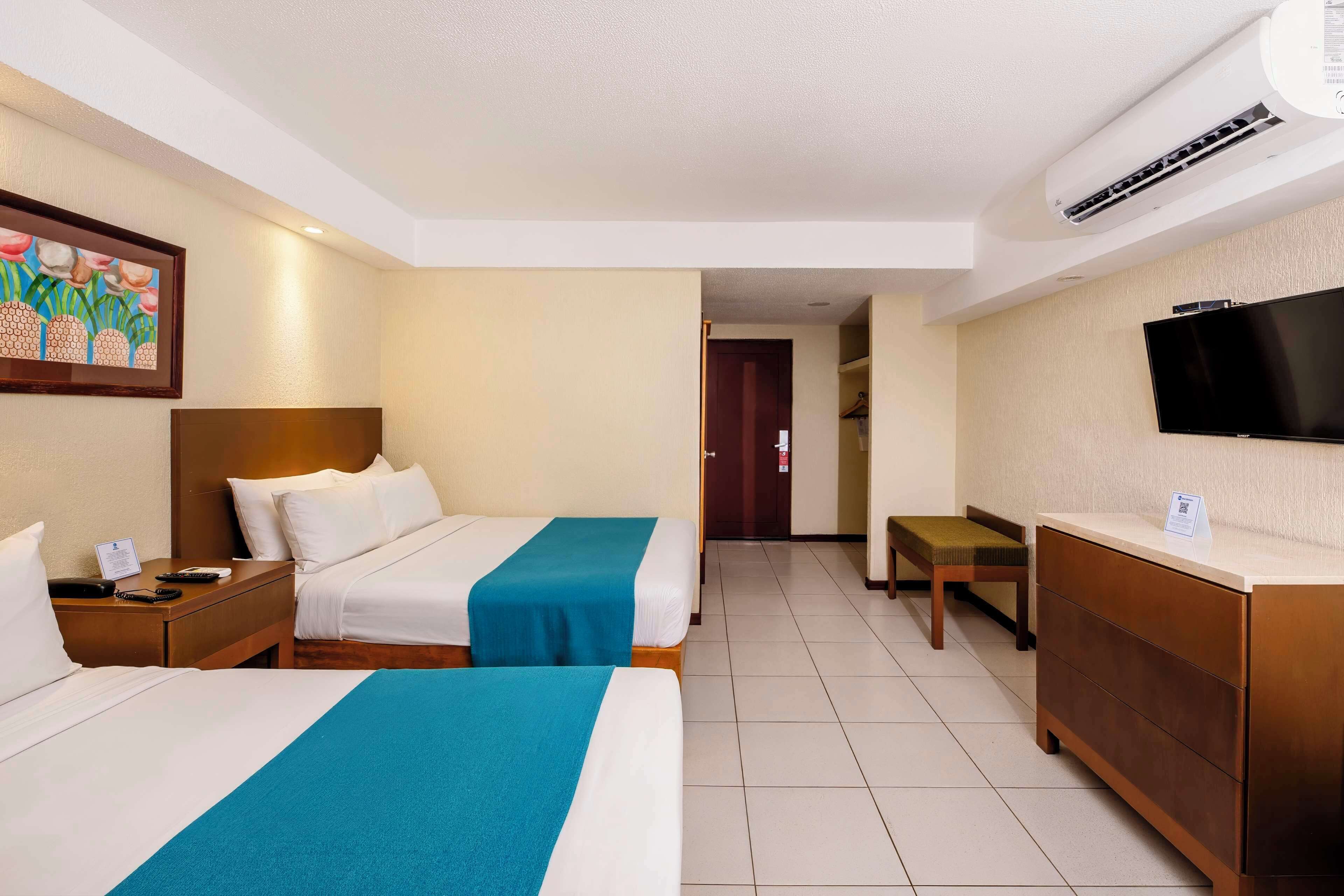 Quarto de hotel com duas camas, uma escrivaninha, uma televisão e um quadro na parede.