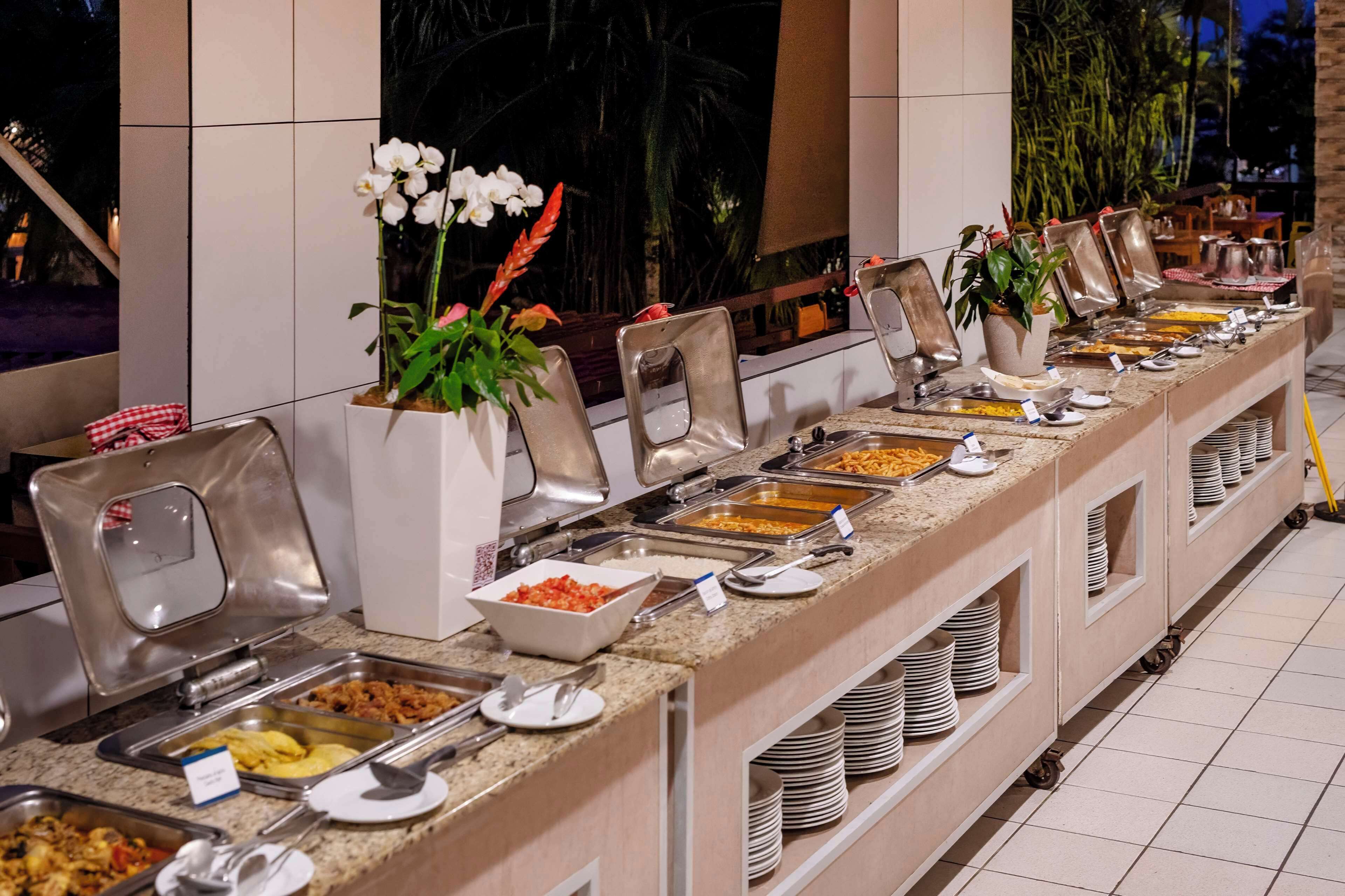 A buffet setup with various food stations, including a salad bar and a pasta bar, with seating arrangements and potted plants.