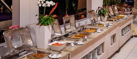 A buffet setup with various food stations, including a salad bar and a pasta bar, with seating arrangements and potted plants.