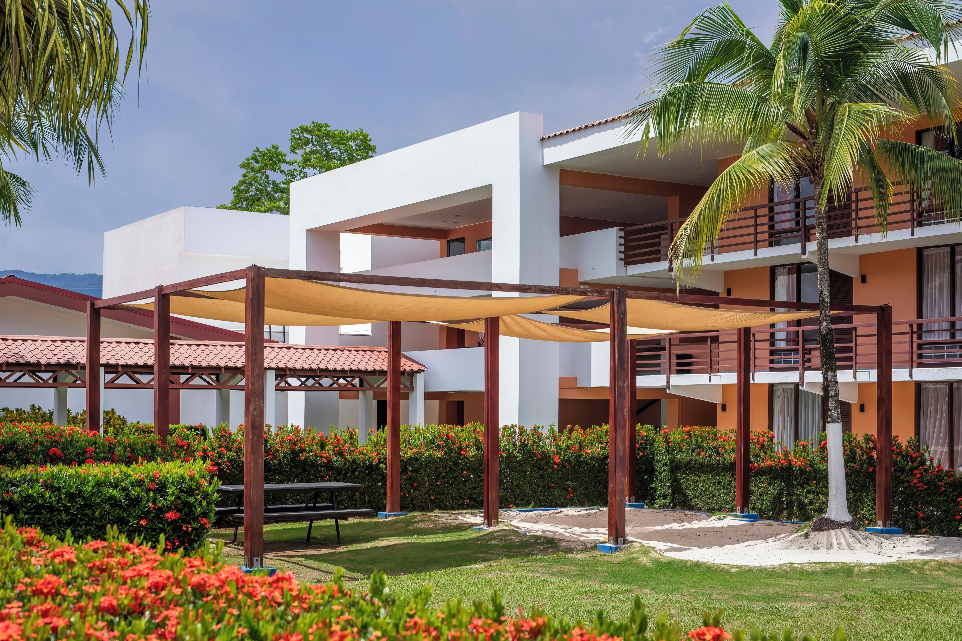 A modern hotel with a covered outdoor area, palm trees, and red flowers.