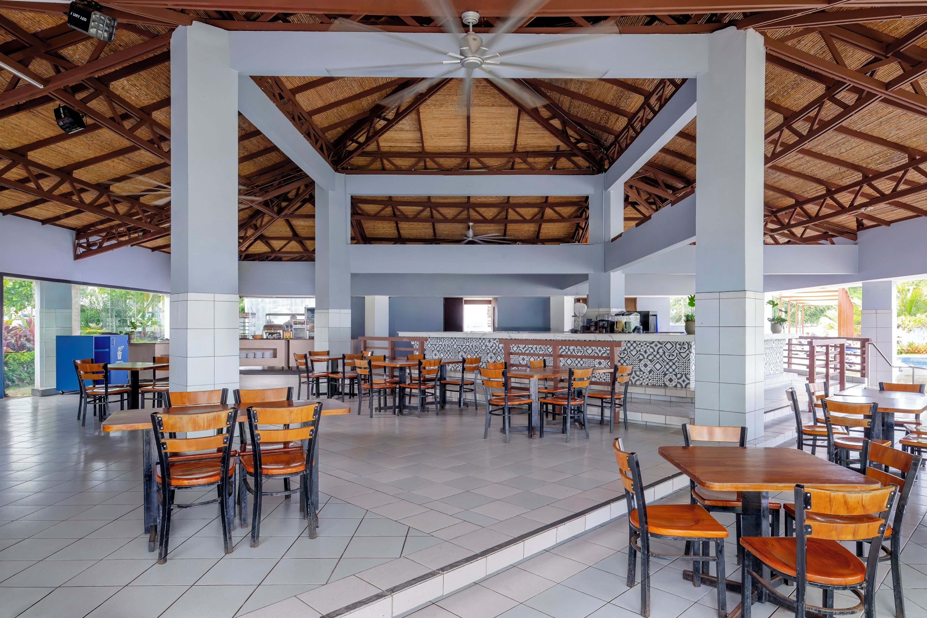 A spacious indoor dining area with multiple wooden tables and chairs, a central kitchen area, and a ceiling fan.