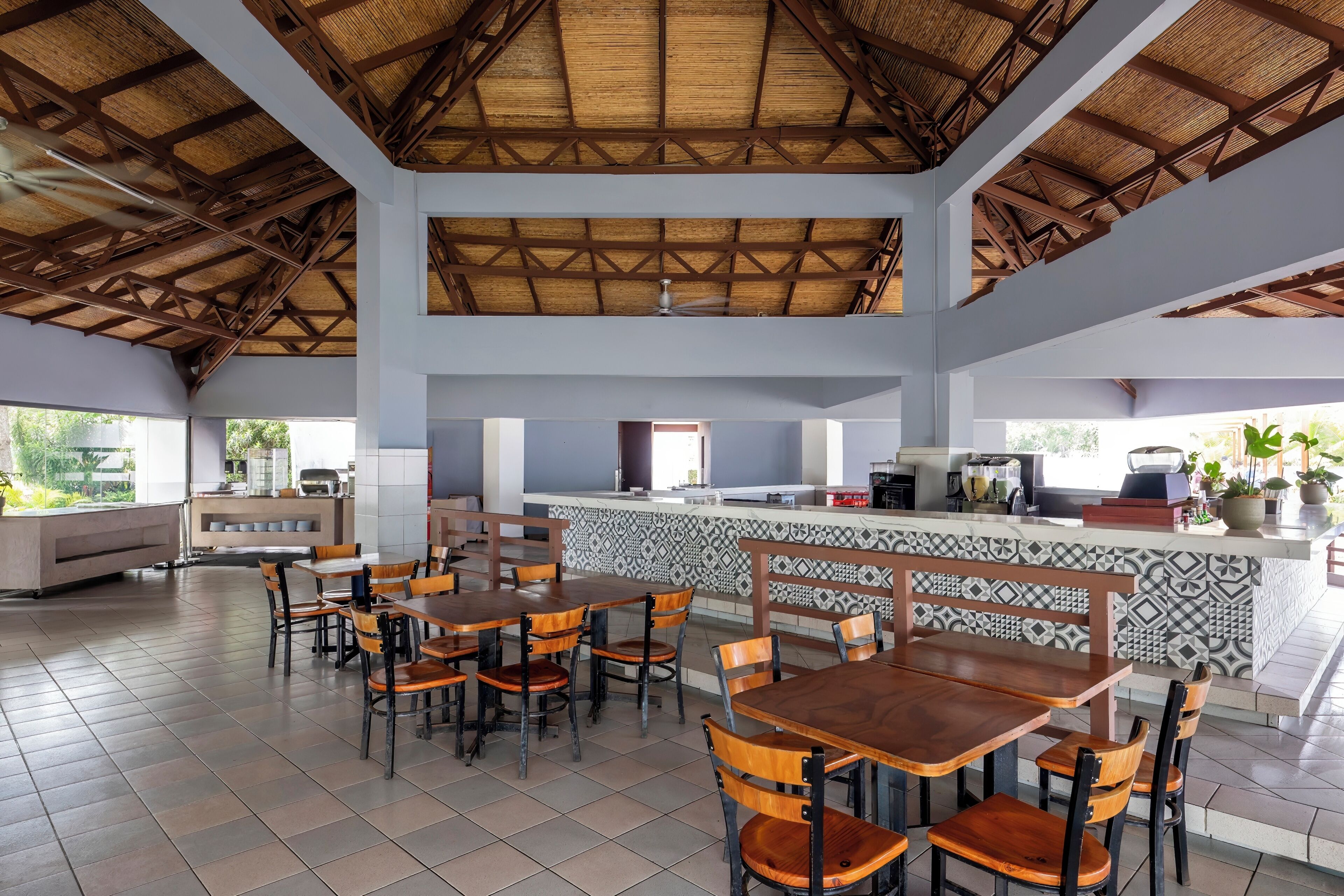 A spacious indoor area with wooden tables and chairs, a counter with a patterned backsplash, and a ceiling with a thatched roof design.