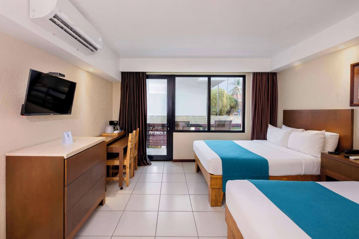 A hotel room with two beds, a desk, a TV, and a balcony with curtains.