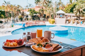 A breakfast table with a variety of food items and a glass of orange juice, set against a backdrop of a swimming pool and palm trees. - Best Western Jaco Beach All-Inclusive Resort (Jacó)
