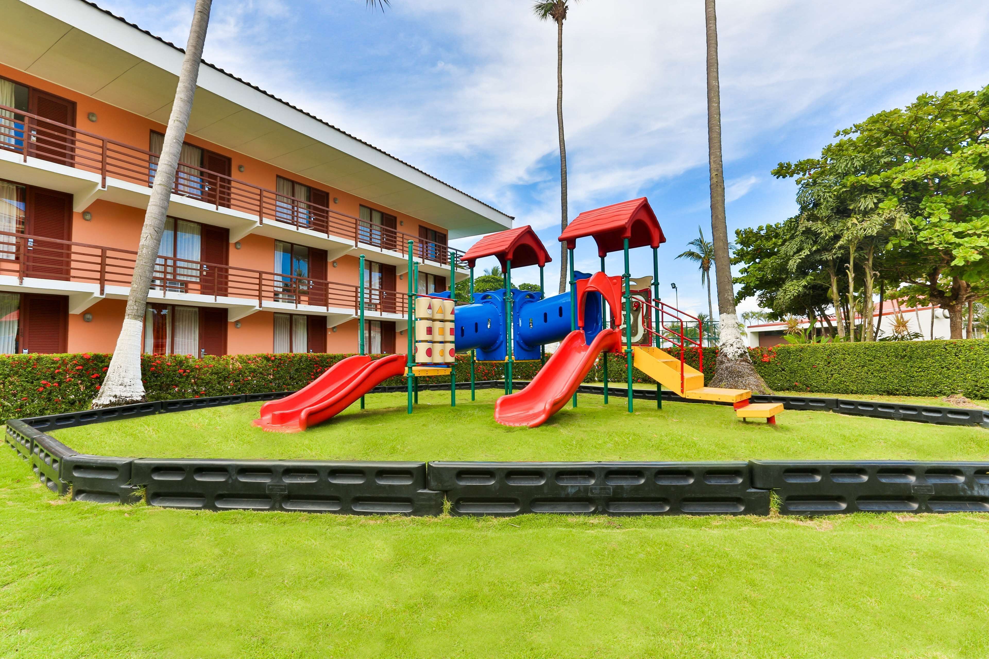 A playground with slides and climbing structures, surrounded by a well-maintained lawn and a building with balconies.
