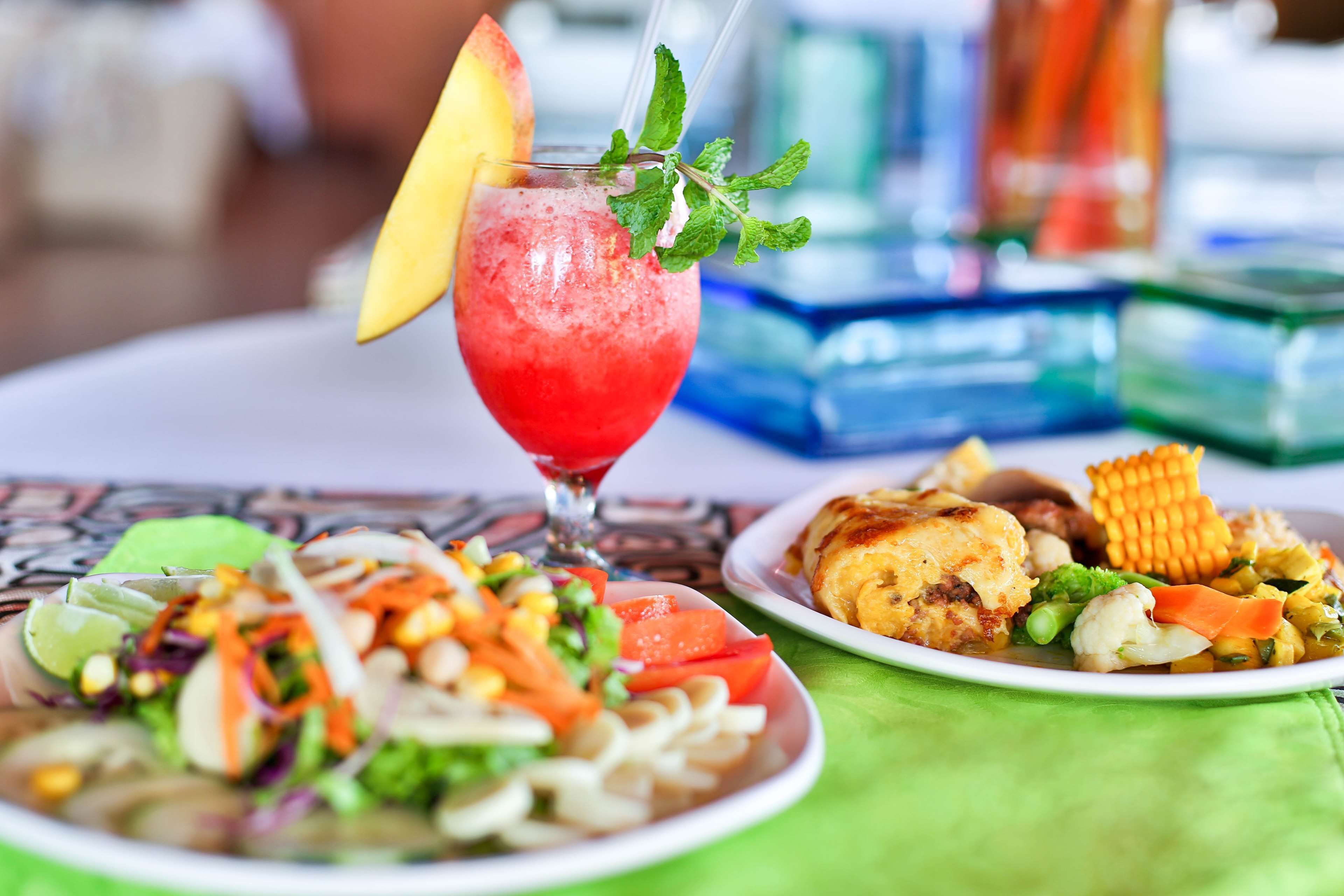 Una mesa con una comida que incluye una ensalada, un plato con maíz y verduras, y un vaso de bebida roja adornado con una rodaja de fruta y hojas de menta.