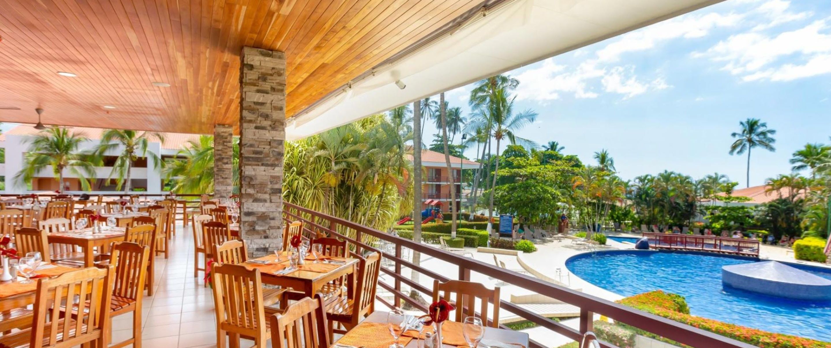A restaurant with wooden tables and chairs, overlooking a pool and palm trees.