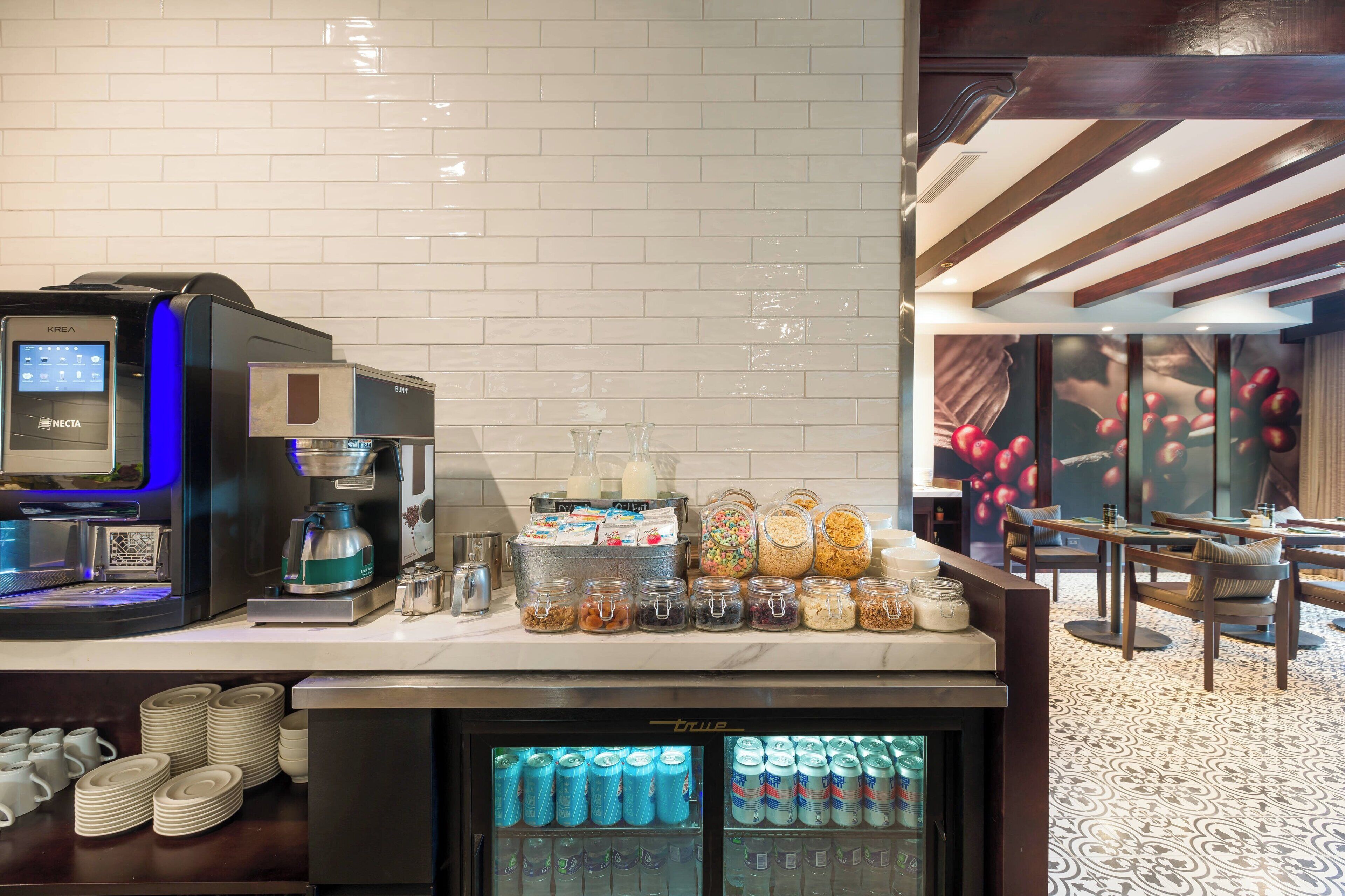 Uma estação de café com uma máquina de café KREa, um dispensador de bebidas e um refrigerador para bebidas.