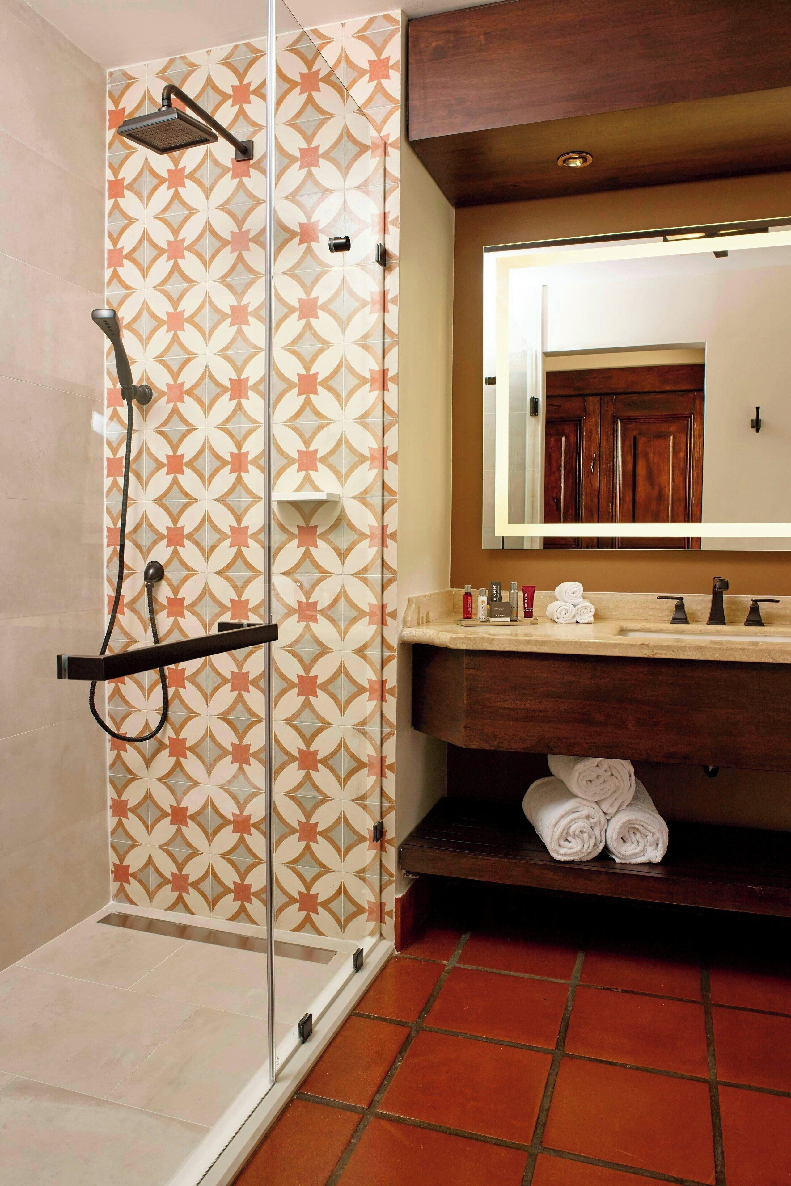 Um banheiro com parede de azulejos estampados, chuveiro com box de vidro, toucador de madeira e espelho sobre a pia.