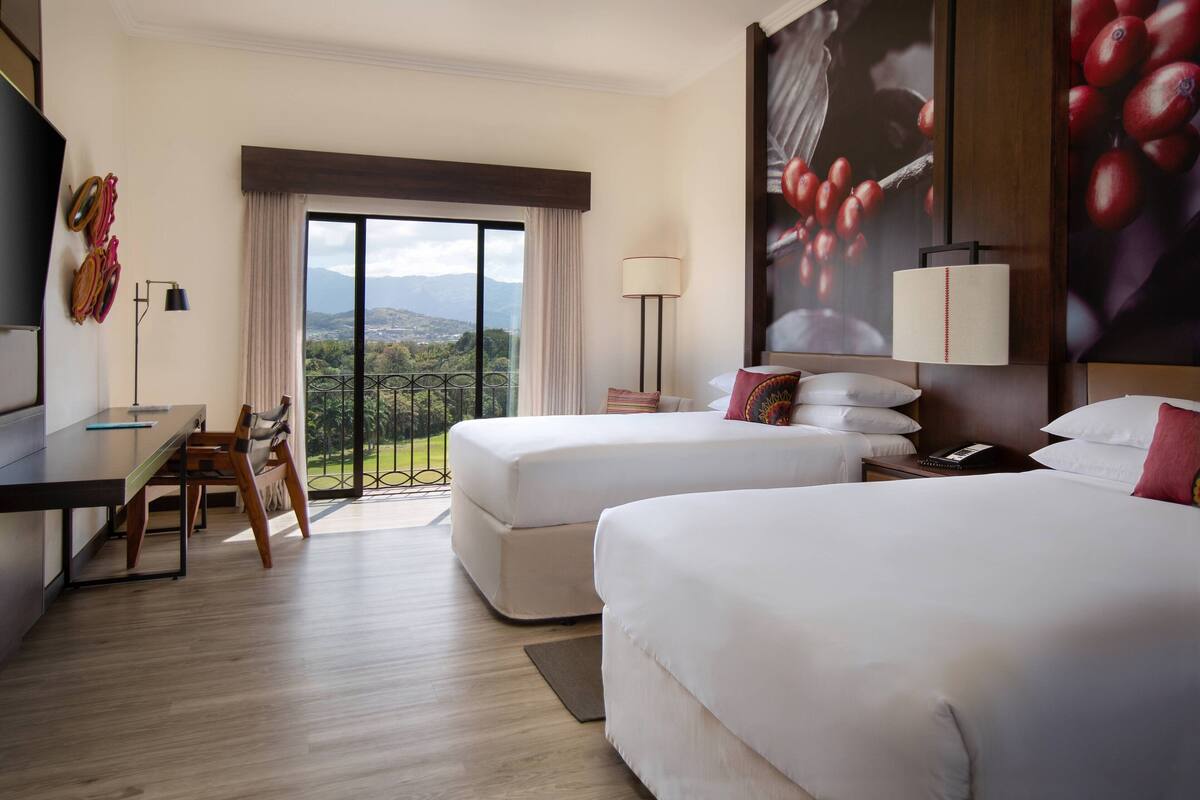 Ein Hotelzimmer mit zwei Betten, einem Schreibtisch und einem Stuhl. Ein großes Fenster bietet Blick auf Berge und Grünflächen.