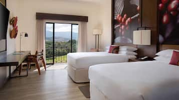 A hotel room with two beds, a desk, and a chair. There is a large window with a view of mountains and greenery.