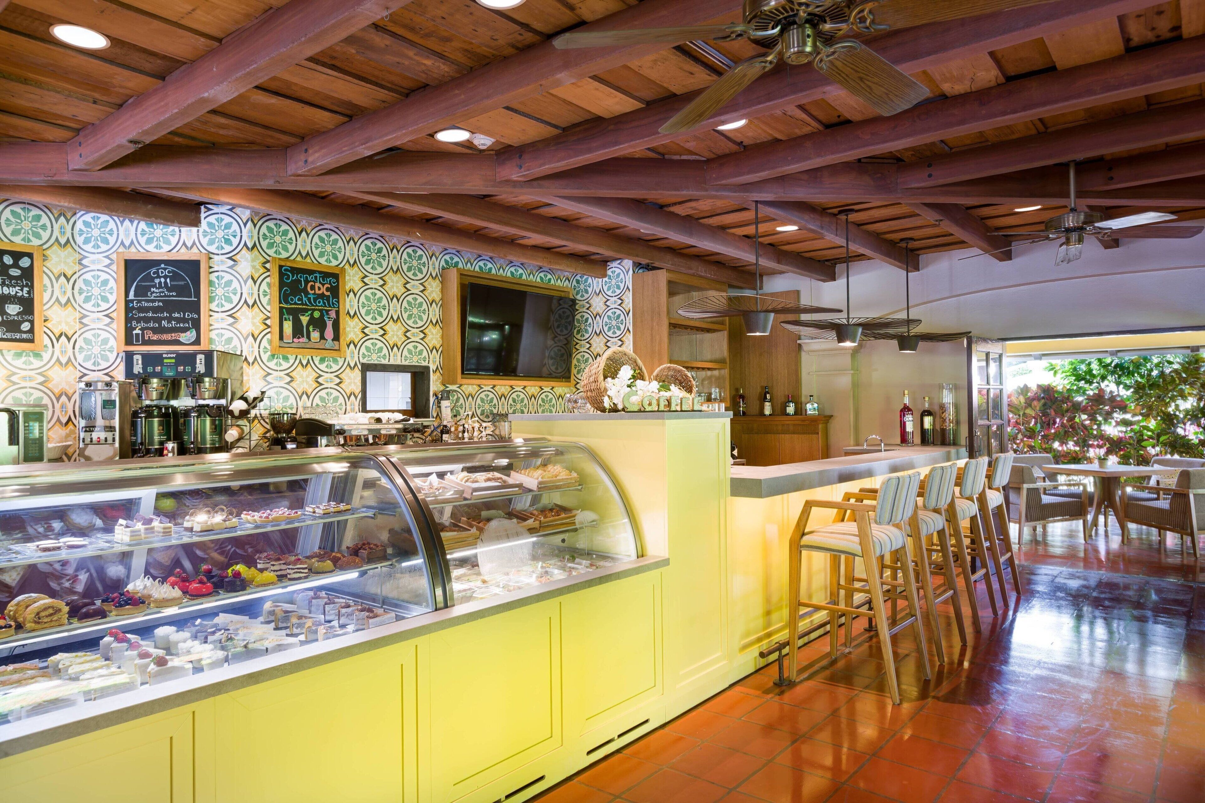 A café with a counter displaying pastries and a selection of drinks. There are wooden beams on the ceiling and a tiled wall with a decorative pattern.