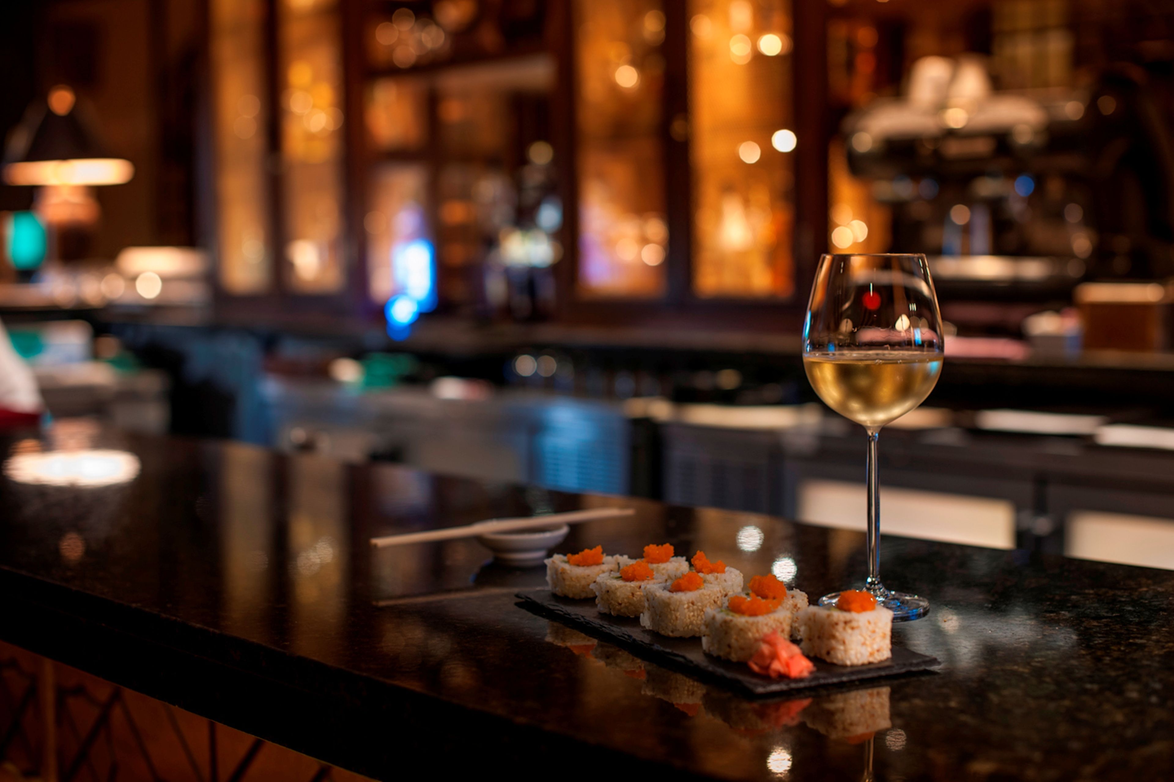 A bar with sushi appetizers and a glass of white wine on a reflective surface.