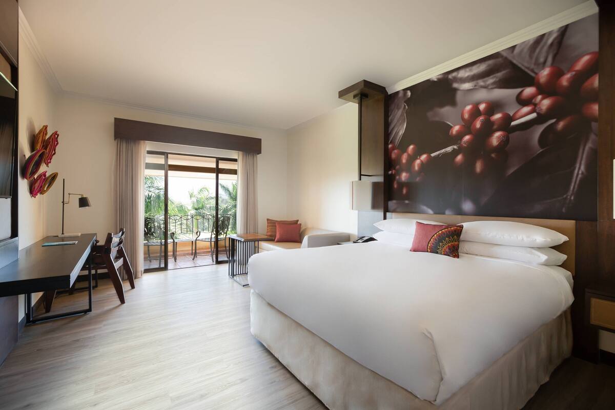 Une chambre d’hôtel avec un grand lit, un bureau et un balcon donnant sur un paysage verdoyant.