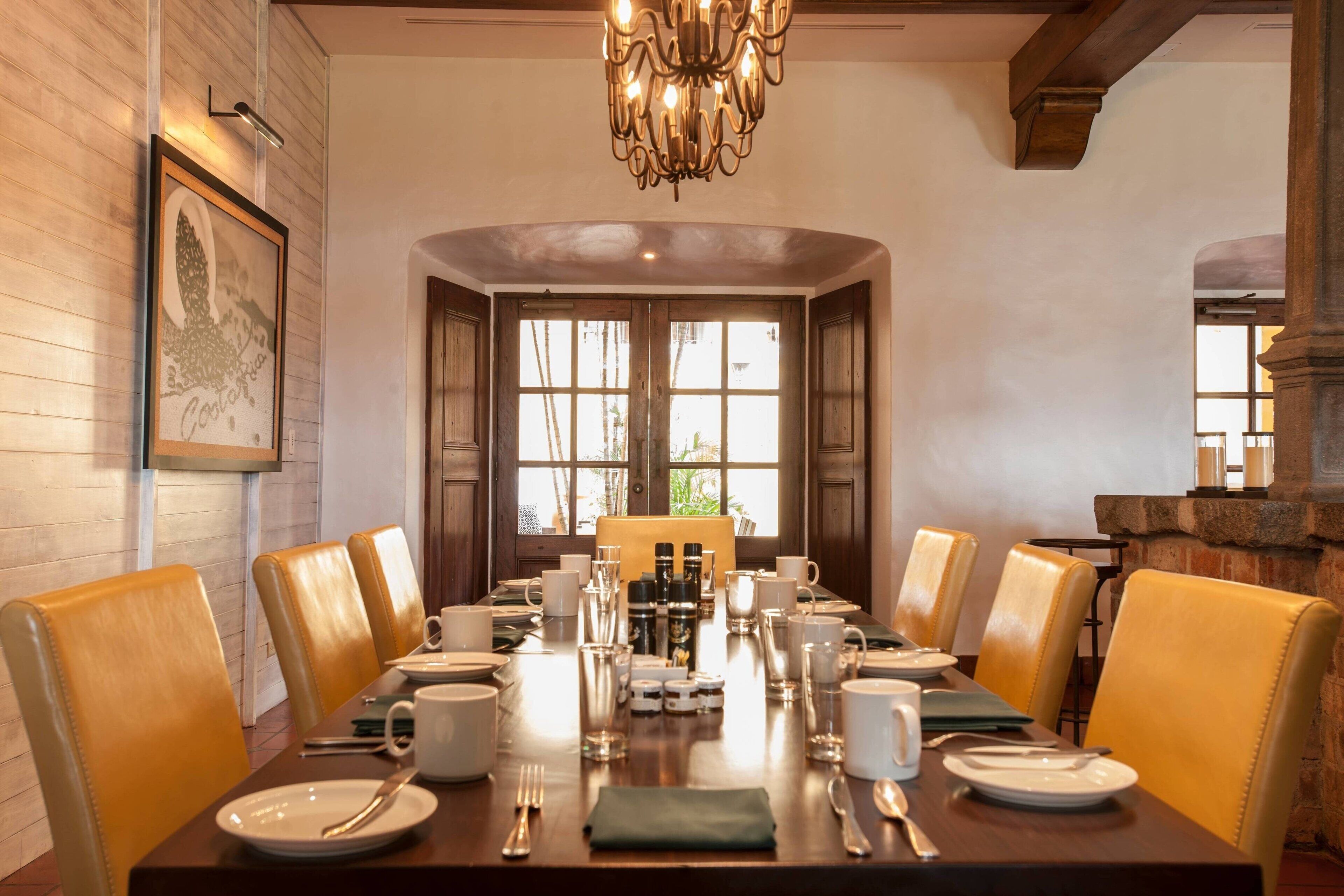 A dining area with a long table set for six, surrounded by leather chairs, with a chandelier above and a framed picture on the wall.