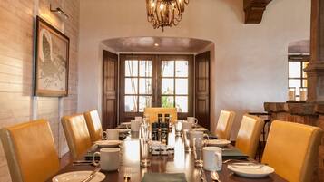 A dining area with a long table set for six, surrounded by leather chairs, with a chandelier above and a framed picture on the wall.