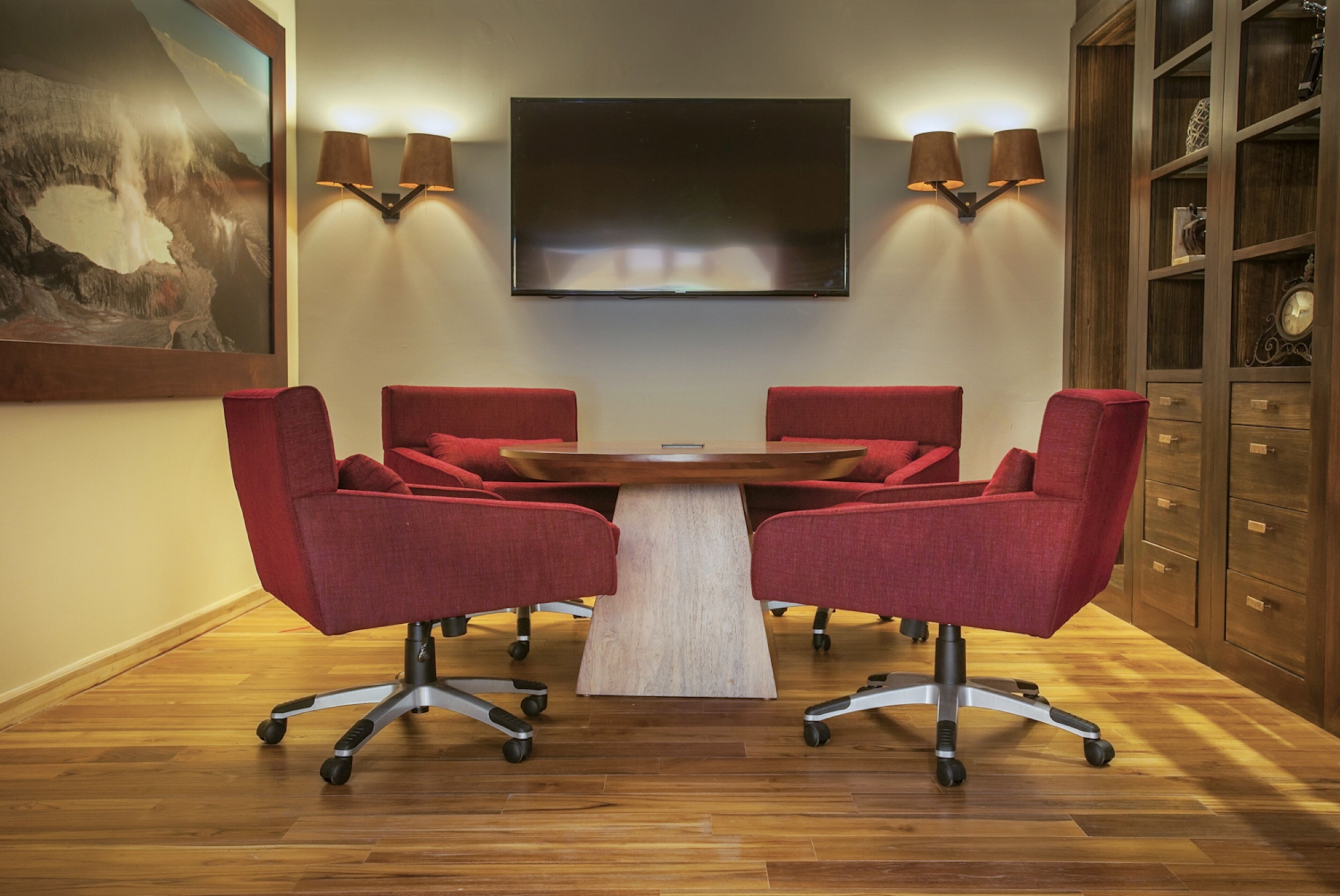 Uma sala de conferências com uma mesa redonda, quatro cadeiras vermelhas, uma TV de tela plana fixada na parede e duas luminárias também fixadas na parede.