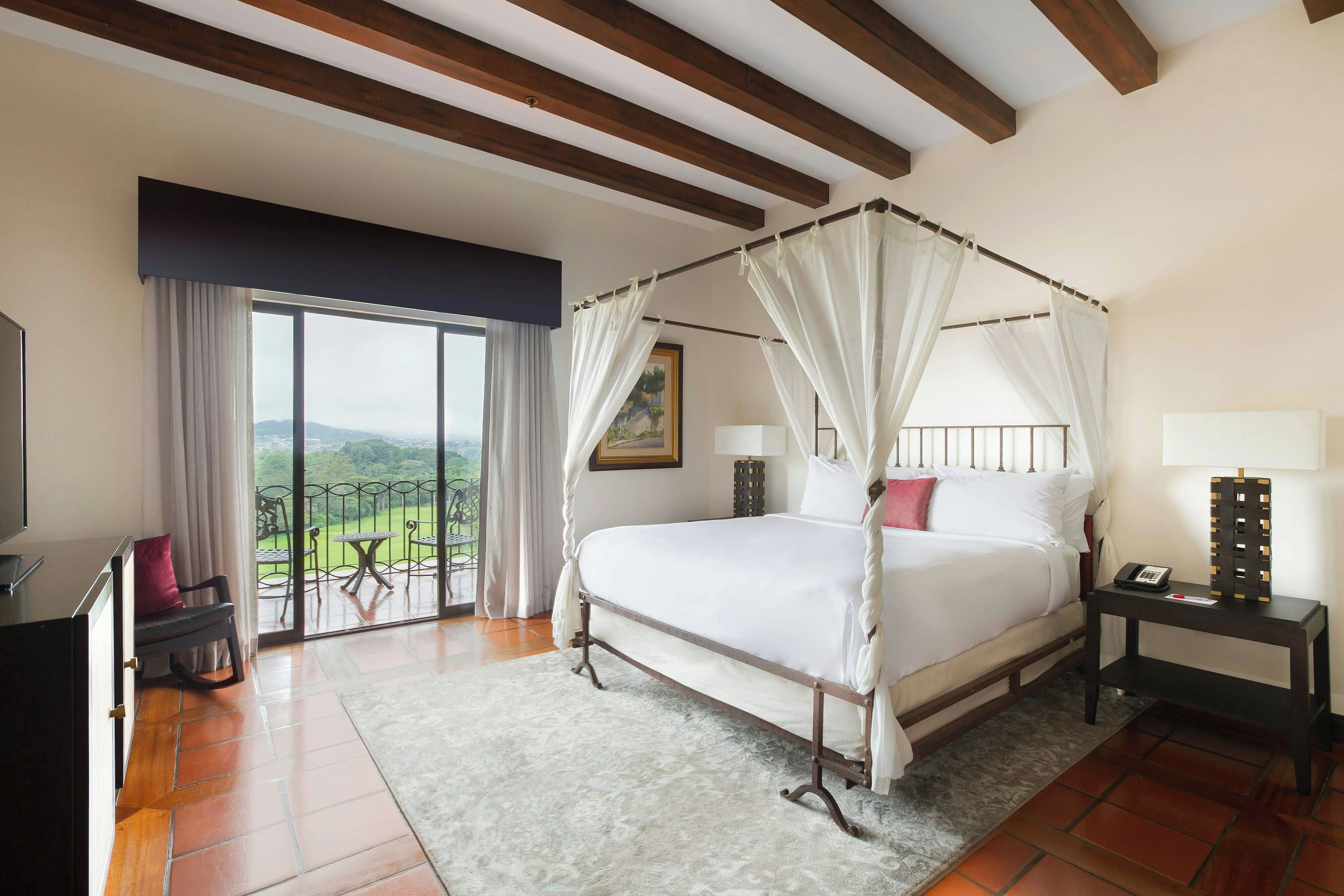 A hotel room with a four-poster bed, a balcony with a table and chairs, and a view of the landscape.