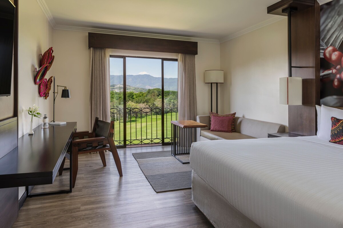 A hotel room with a large bed, a desk, a chair, a sofa, and a balcony with a view of the mountains.