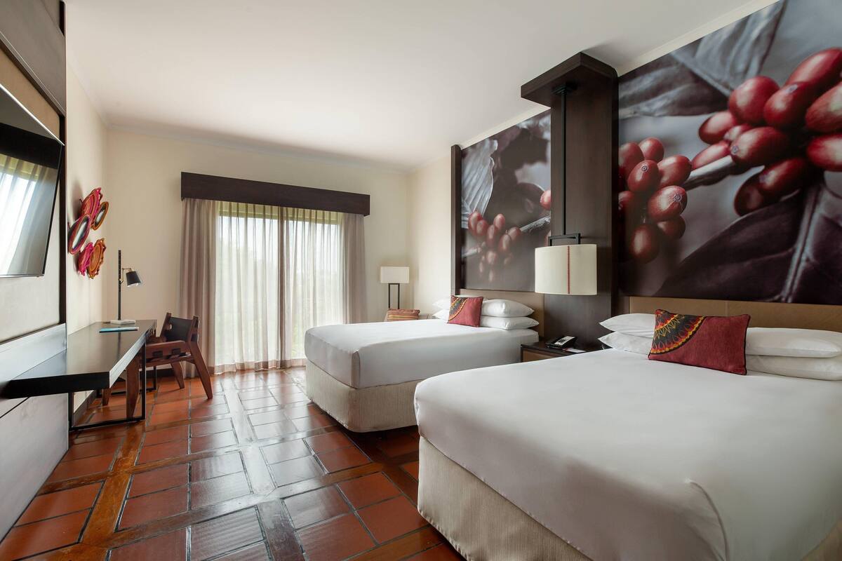Ein Hotelzimmer mit zwei Betten, einem Schreibtisch, einem Sessel, einem großen Fenster mit Vorhängen und einem an der Wand befestigten Fernseher.