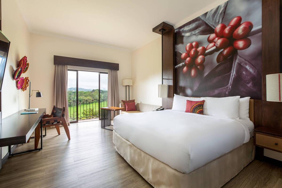Une chambre d’hôtel avec un grand lit, un bureau, une chaise et un balcon donnant sur un paysage verdoyant.
