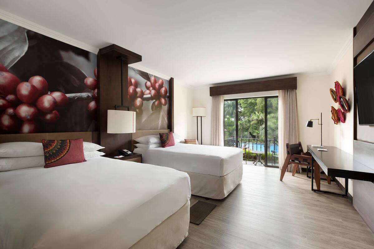 Ein Hotelzimmer mit zwei Betten, einem Schreibtisch, einem Sessel, einem Fernseher und einem großen Fenster mit Blick auf einen Pool.
