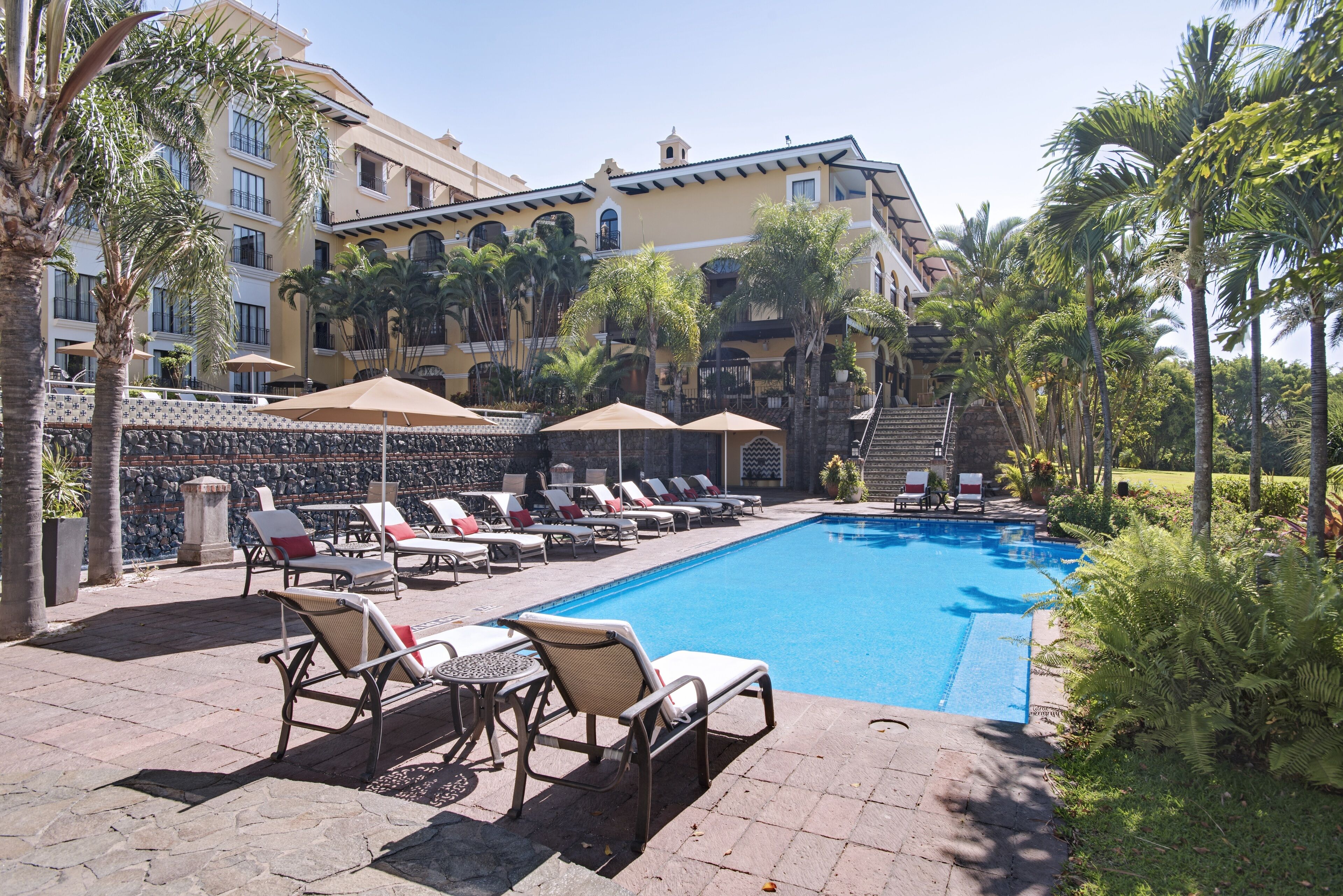 A poolside area with lounge chairs and umbrellas, surrounded by palm trees and a building with balconies.