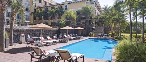 A poolside area with lounge chairs and umbrellas, surrounded by palm trees and a building with balconies.