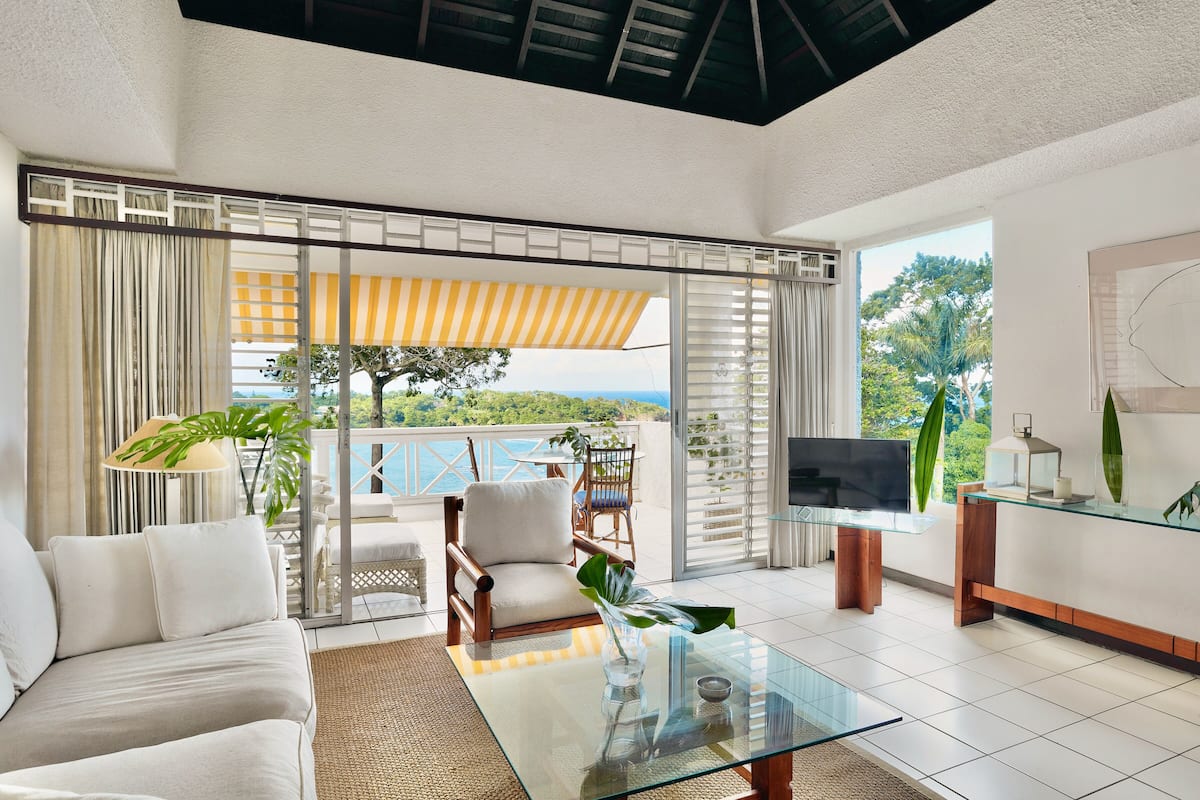 A modern living room with a sofa, armchair, and glass coffee table, a TV, and a balcony with a view.