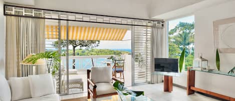 A modern living room with a sofa, armchair, and glass coffee table, a TV, and a balcony with a view.