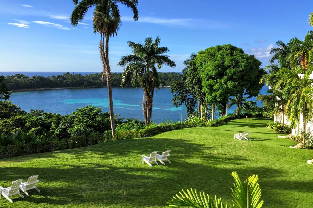 A lush, green lawn with white lawn chairs and a view of a calm sea and palm trees.