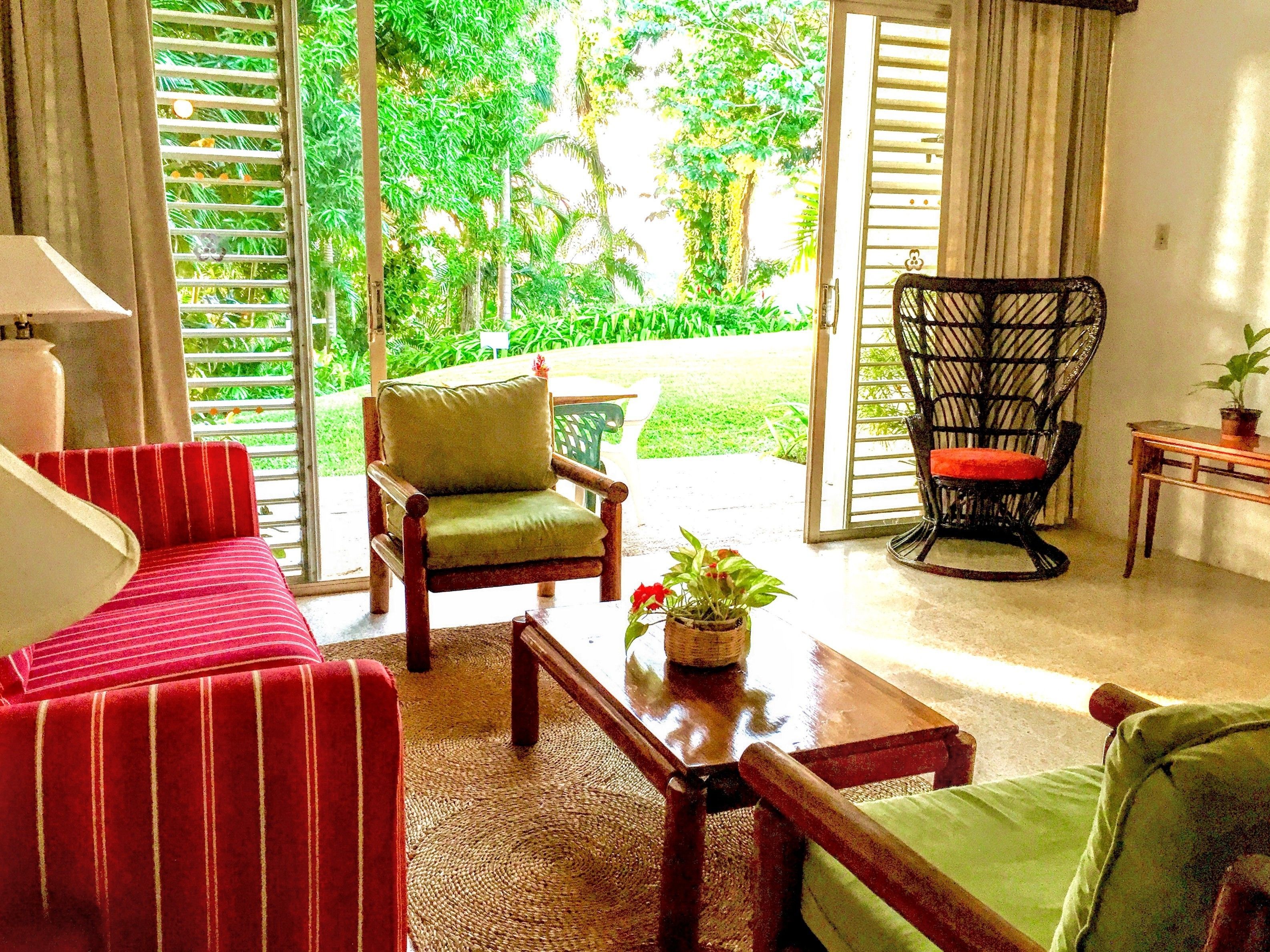 A room with a red-striped sofa, a green armchair, and a wicker chair with a red cushion. There is a small table with a potted plant.