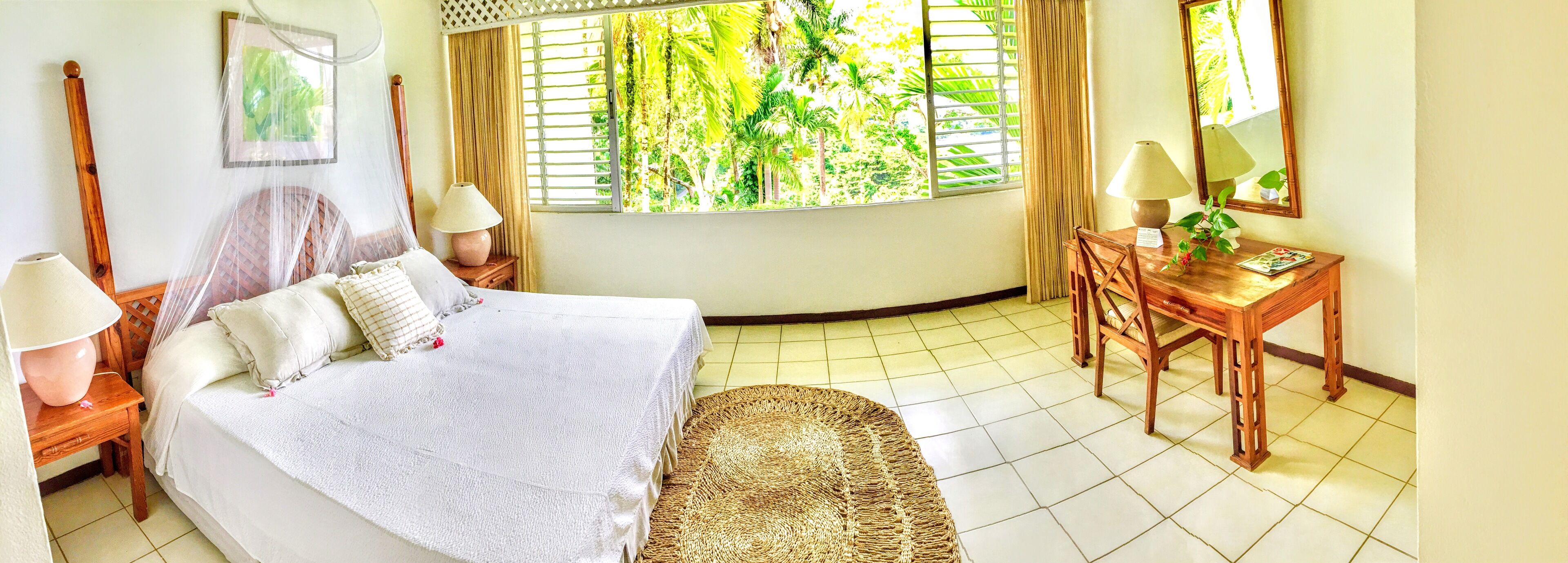 A bedroom with a bed, bedside tables, a desk, a chair, a window with a view of greenery, and a mirror.