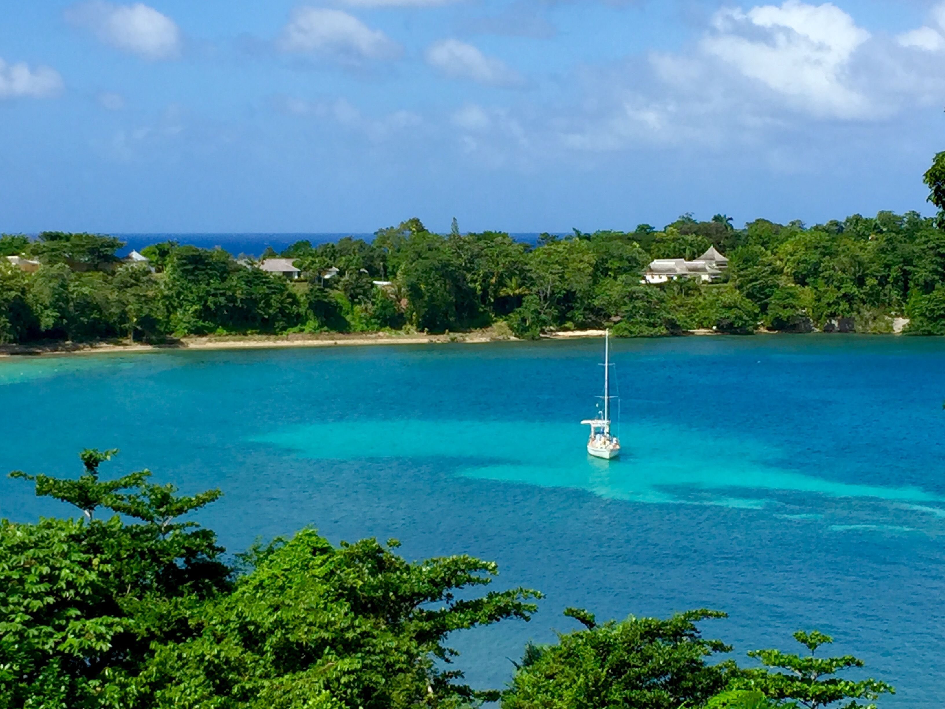 A clear view of a turquoise sea with a sailboat, surrounded by lush greenery and a few buildings in the distance.