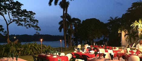 Un espace de restauration en plein air, avec des tables dressées pour le repas, entouré de palmiers et offrant une vue sur l’eau.