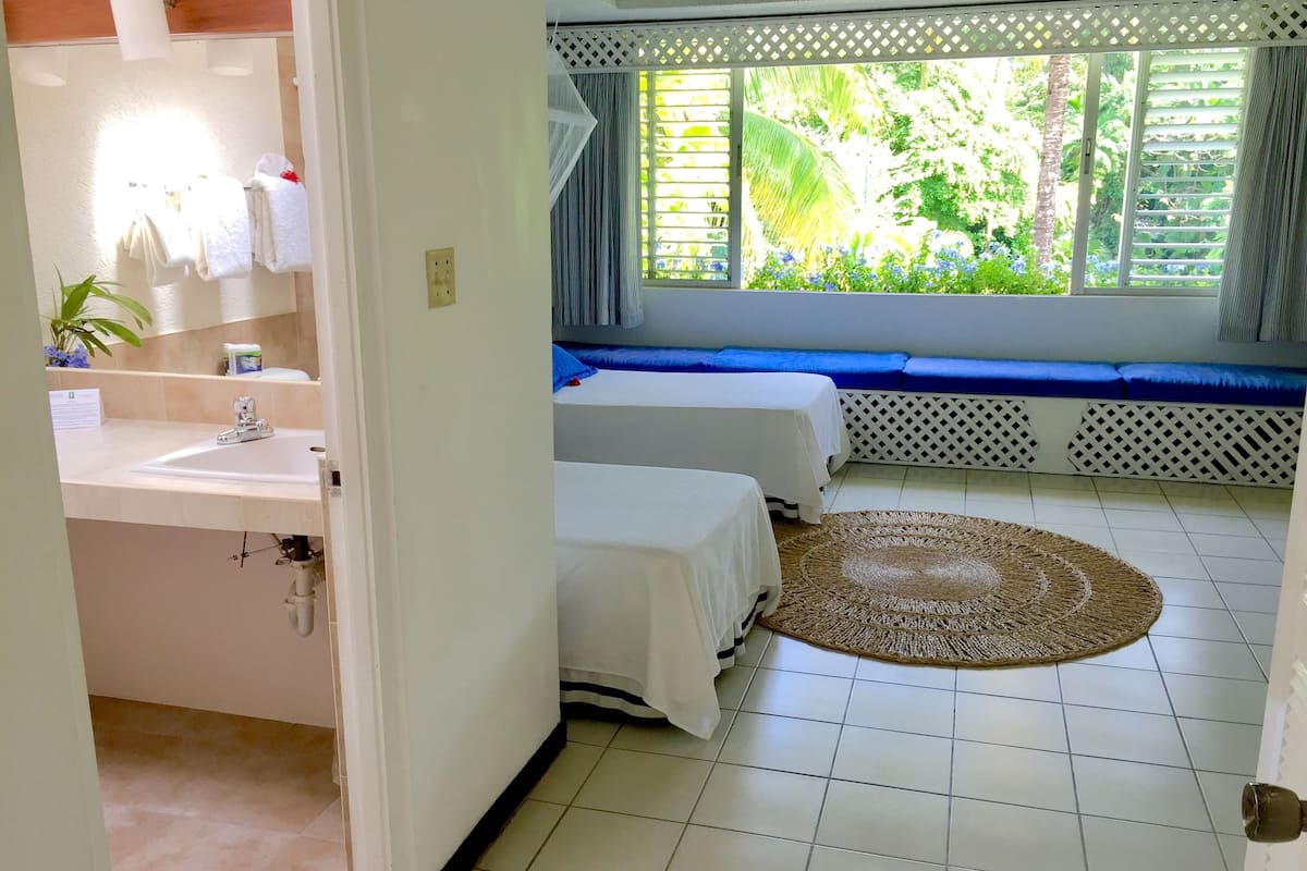 A hotel room with a bathroom, two beds, a window with a view of greenery, and a woven rug.