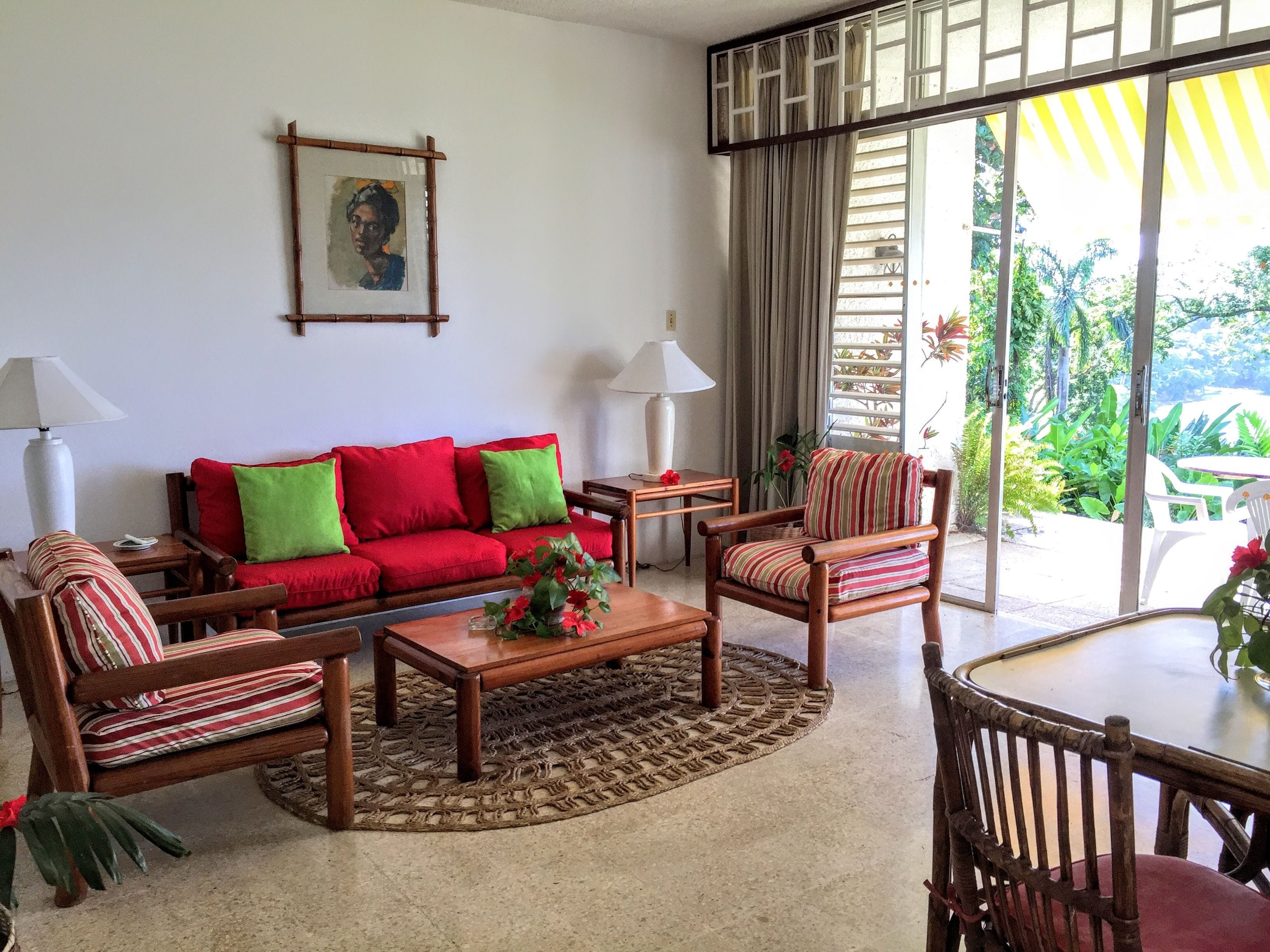 A living room with a red sofa, striped armchairs, a coffee table, and a dining table. There is a painting on the wall and a view of greenery through the window.