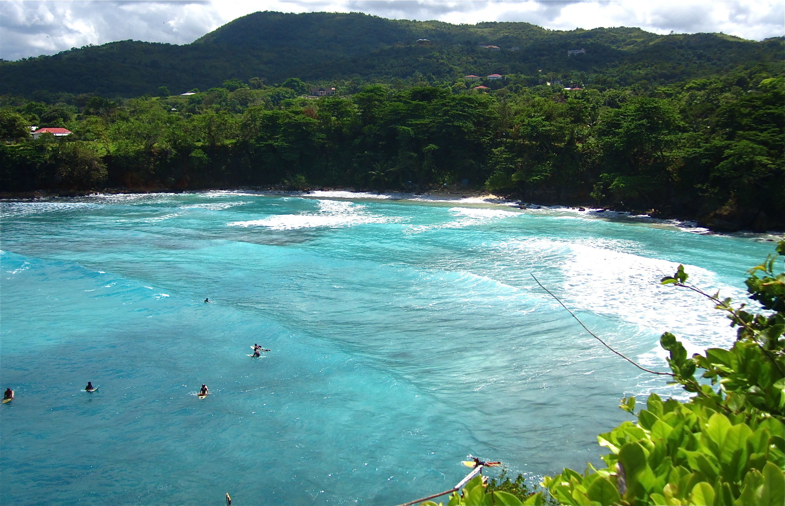 Eine Küstenlandschaft mit klarem türkisblauem Wasser, Surfern und üppigen, grünen Hügeln im Hintergrund.