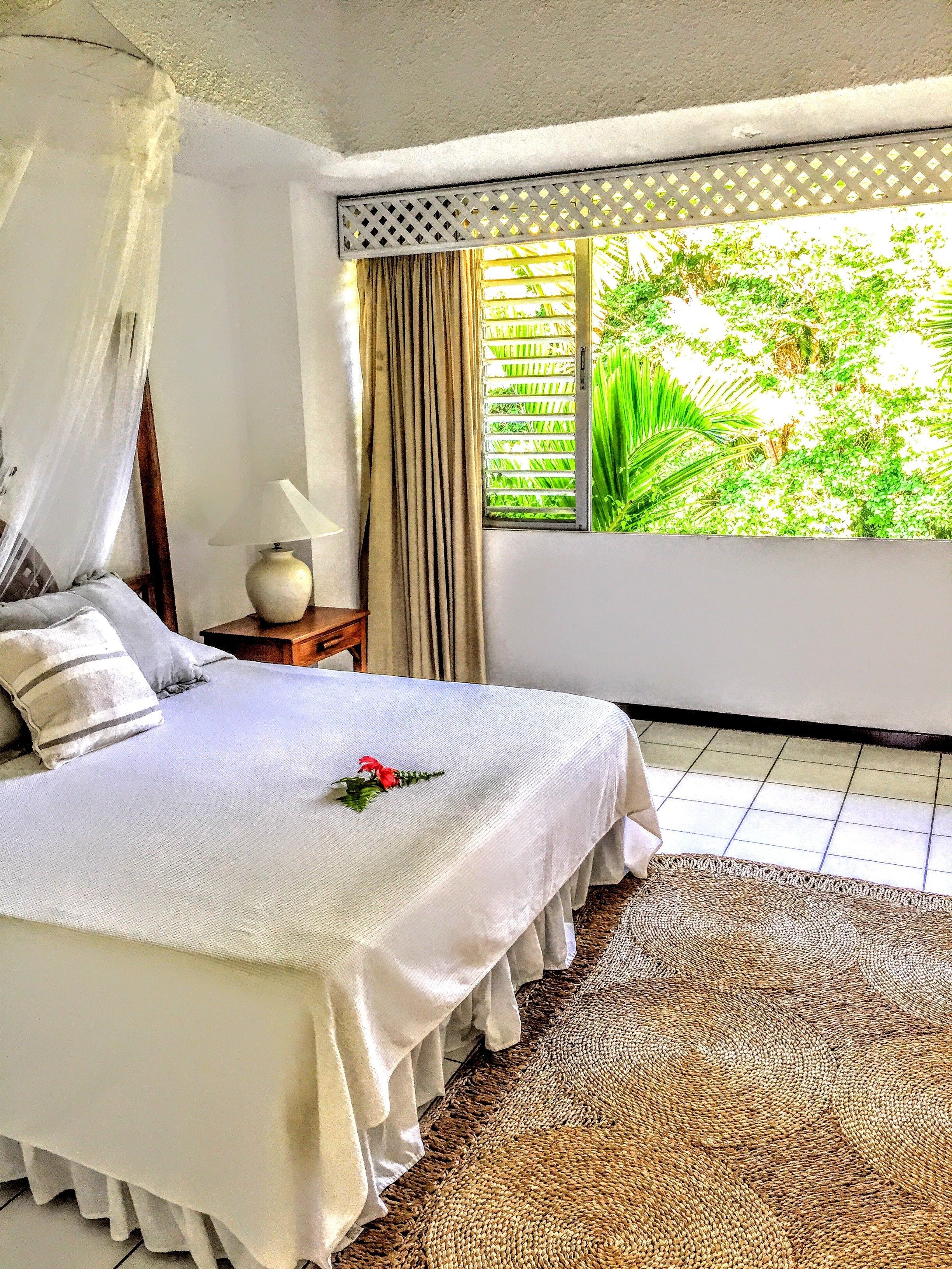 A bedroom with a bed, a nightstand, a lamp, and a window with a view of greenery.