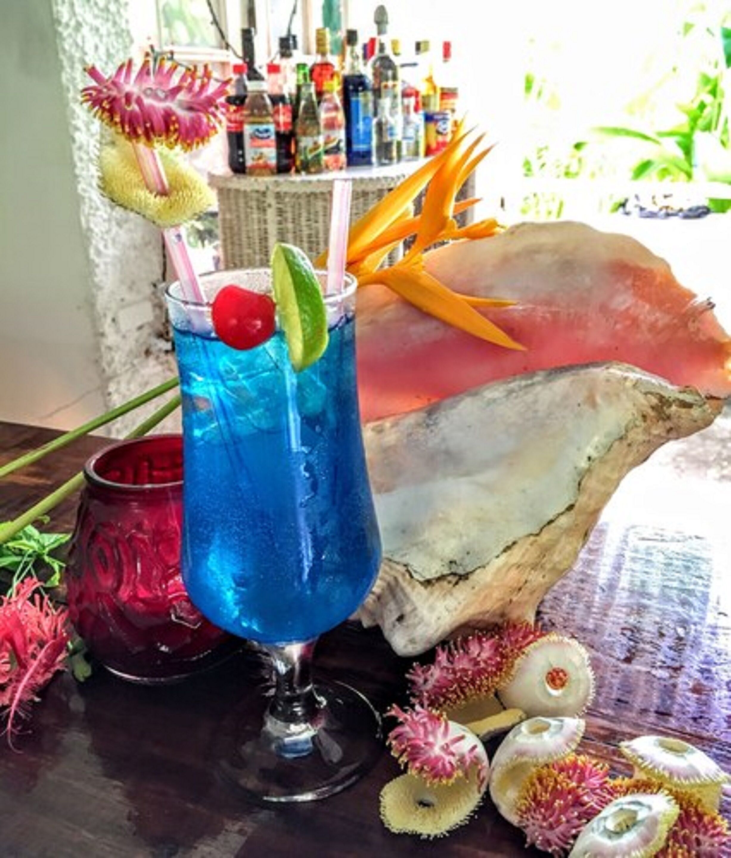 A cocktail glass with a blue drink, garnished with a cherry and lime, on a table with decorative shells and a red mug.